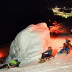 Menschen fahren nachts mit Holzschlitten einen verschneiten Hang hinunter, beleuchtet von orangefarbenen Lichtern, vorbei an einem großen schneebedeckten Felsblock.
