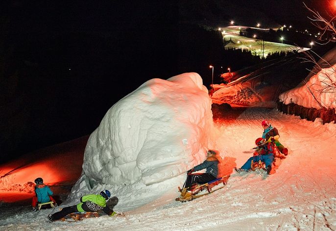 Menschen fahren nachts mit Holzschlitten einen verschneiten Hang hinunter, beleuchtet von orangefarbenen Lichtern, vorbei an einem großen schneebedeckten Felsblock.
