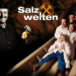 Eine Gruppe von Menschen in weißen Anzügen gleitet in einem Salzbergwerk eine Holzrutsche hinunter. Ein Bergmann hält eine Laterne und den Text „Salzwelten Faszination“ in der Hand.