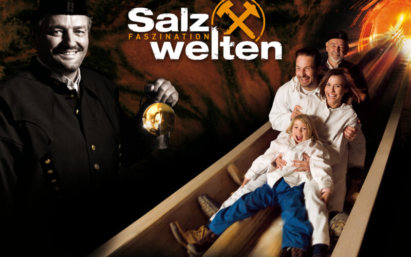 Eine Gruppe von Menschen in weißen Anzügen gleitet in einem Salzbergwerk eine Holzrutsche hinunter. Ein Bergmann hält eine Laterne und den Text „Salzwelten Faszination“ in der Hand.