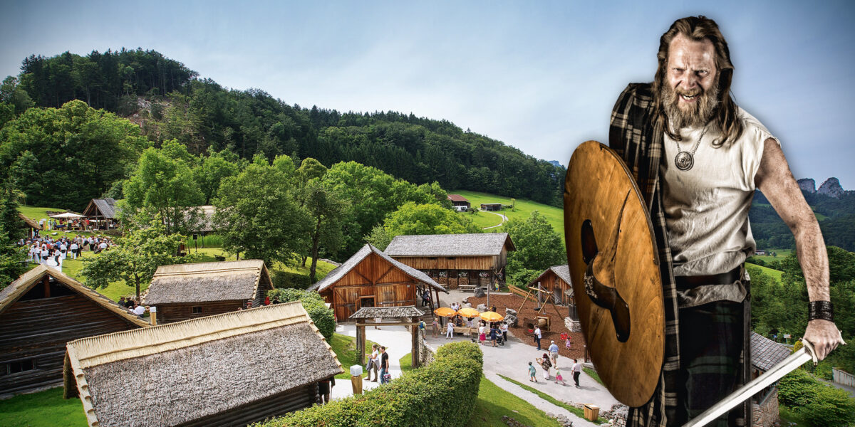 Ein als Wikinger gekleideter Mann mit Schild und Schwert steht vor einem rekonstruierten historischen Dorf mit Holzhäusern und Besuchern.