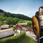 Ein als Wikinger gekleideter Mann mit Schild und Schwert steht vor einem rekonstruierten historischen Dorf mit Holzhäusern und Besuchern.