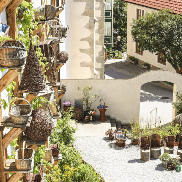 Im Freien stehen geflochtene Körbe auf einem Holzgestell neben einer Steinmauer, weitere Körbe und Pflanzen sind in einem sonnigen Innenhof aufgestellt.