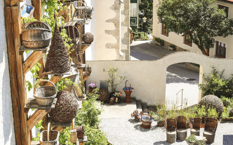 Im Freien stehen geflochtene Körbe auf einem Holzgestell neben einer Steinmauer, weitere Körbe und Pflanzen sind in einem sonnigen Innenhof aufgestellt.