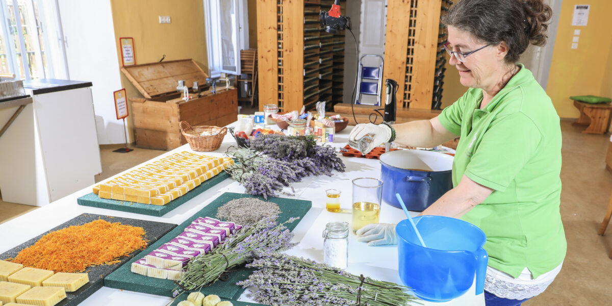 Eine Frau in einem grünen Hemd stellt handgemachte Seifen her und arrangiert Trockenblumen auf einem Tisch in einer hellen, organisierten Werkstatt.