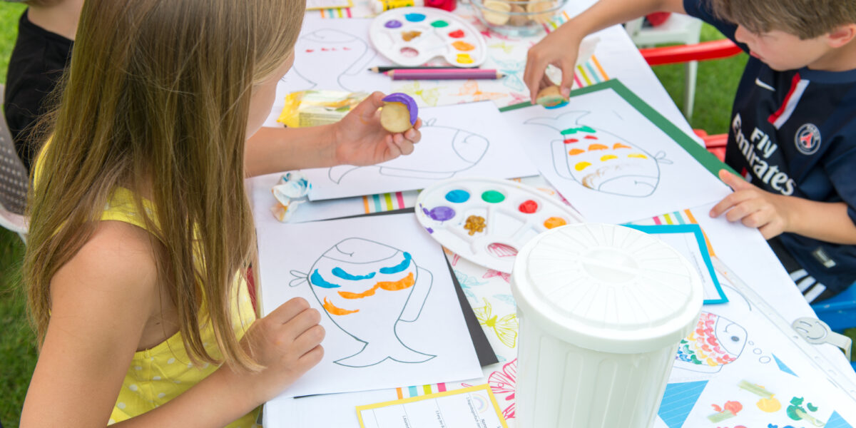 Kinder malen mit bunten Farben Fischumrisse auf Papier. Sie sitzen an einem Tisch, umgeben von Malutensilien und Farbpaletten.