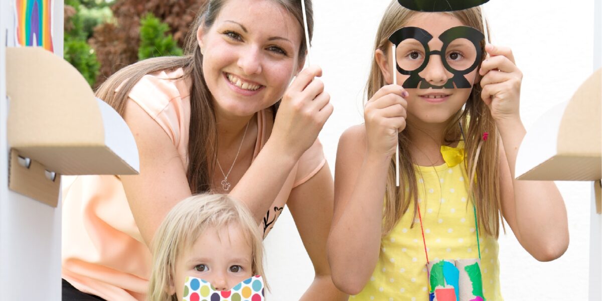 Drei Kinder mit bunten Papierhüten und Brillen an einem Spielmarktstand, die lächeln und mit ausgestelltem Spielzeuggemüse für die Kamera posieren.
