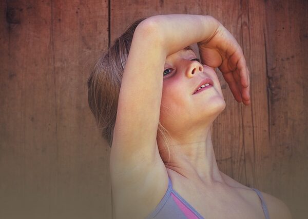 Ein junges Mädchen steht vor einer Holzwand mit erhobenem rechten Arm, der einen Teil ihrer Stirn und ihrer Augen bedeckt.