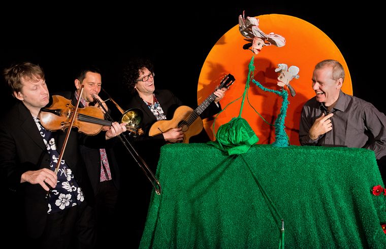 Vier Männer treten auf; zwei spielen Instrumente (Geige, Posaune), einer spielt Gitarre, und einer interagiert mit zwei Puppen auf einer grünen Bühne mit orangefarbenem Hintergrund.