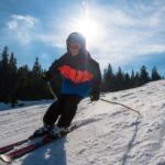 Ein Skifahrer mit Helm und Winterausrüstung fährt eine verschneite Piste hinunter, im Hintergrund ein anderer Skifahrer und Bäume in der strahlenden Sonne.