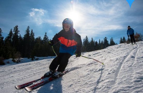 Ein Skifahrer mit Helm und Winterausrüstung fährt eine verschneite Piste hinunter, im Hintergrund ein anderer Skifahrer und Bäume in der strahlenden Sonne.