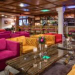 Farbenfroher Lounge-Bereich mit rosafarbenen und gelben Sesseln, gläsernen Couchtischen und einer Bar im Hintergrund, deren Decke und Wände holzvertäfelt sind.