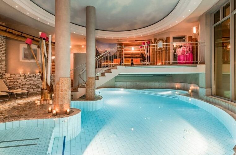 Hallenbad mit geschwungenen Rändern, umgeben von modernem Dekor, Kerzen, Liegestühlen, einer Treppe und stimmungsvoller Beleuchtung.