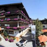 Ein mehrstöckiges Alpenhotel mit Holzbalkonen, die mit violetten Blumen geschmückt sind, mit Sitzgelegenheiten im Freien und einem Cabrio, das an einem sonnigen Tag in der Nähe parkt.