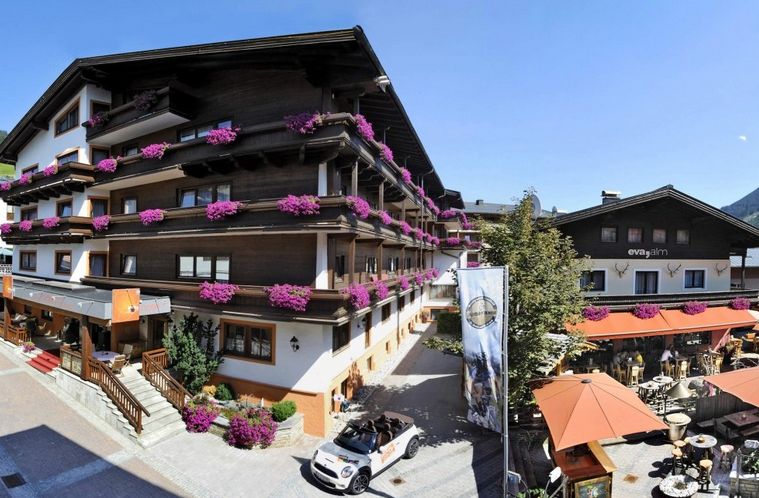 Ein mehrstöckiges Alpenhotel mit Holzbalkonen, die mit violetten Blumen geschmückt sind, mit Sitzgelegenheiten im Freien und einem Cabrio, das an einem sonnigen Tag in der Nähe parkt.