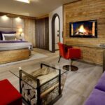 Modernes Hotelzimmer mit einem Doppelbett, holzgetäfelten Wänden, einem Kamin an der Wand, einem gläsernen Couchtisch mit Brennholz, einem roten Stuhl und einem lila Sofa.