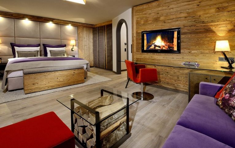 Modernes Hotelzimmer mit einem Doppelbett, holzgetäfelten Wänden, einem Kamin an der Wand, einem gläsernen Couchtisch mit Brennholz, einem roten Stuhl und einem lila Sofa.
