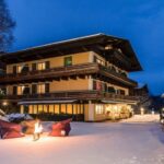 Ein dreistöckiges Gebäude im alpinen Stil mit Balkonen ist in der Abenddämmerung beleuchtet, umgeben von Schnee; ein kleines Feuer mit Sitzgelegenheiten und ein Pavillon stehen im verschneiten Hof.