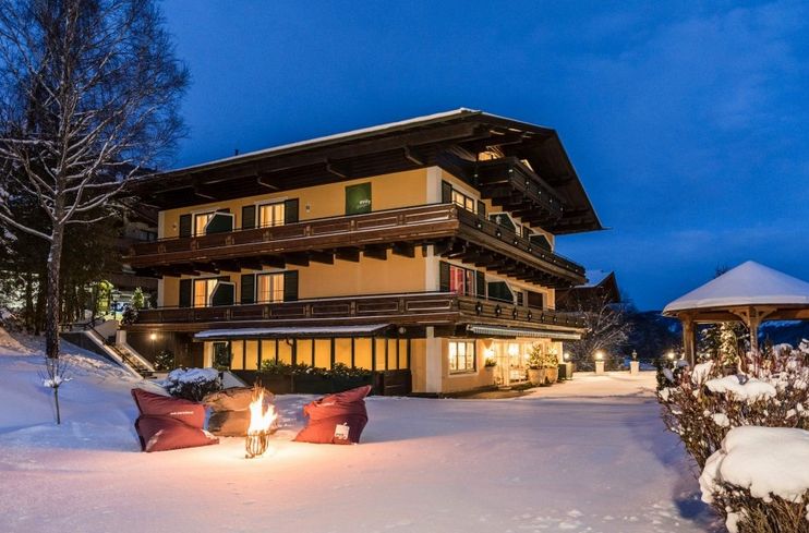 Ein dreistöckiges Gebäude im alpinen Stil mit Balkonen ist in der Abenddämmerung beleuchtet, umgeben von Schnee; ein kleines Feuer mit Sitzgelegenheiten und ein Pavillon stehen im verschneiten Hof.