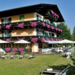 Ein dreistöckiges Hotelgebäude mit blumengeschmückten Balkonen, grünen Fensterläden und Liegestühlen im Freien auf einer Wiese mit Sonnenschirmen und Sitzgelegenheiten im Innenhof.