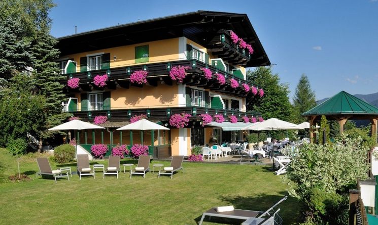 Ein dreistöckiges Hotelgebäude mit blumengeschmückten Balkonen, grünen Fensterläden und Liegestühlen im Freien auf einer Wiese mit Sonnenschirmen und Sitzgelegenheiten im Innenhof.