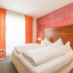 Ein Hotelzimmer mit einem Doppelbett, weißem Bettzeug, einem Kopfteil aus Holz, einer roten Akzentwand, orangefarbenen Vorhängen und einem weißen Sessel am Fenster.