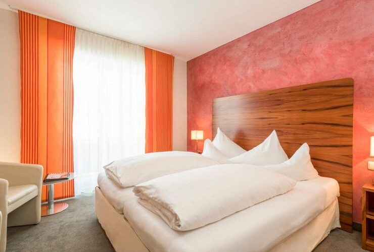 Ein Hotelzimmer mit einem Doppelbett, weißem Bettzeug, einem Kopfteil aus Holz, einer roten Akzentwand, orangefarbenen Vorhängen und einem weißen Sessel am Fenster.