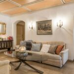 Ein Wohnzimmer mit beigen Sofas, einem Couchtisch aus Glas, gemusterten Kissen, Wandleuchten, gerahmten Kunstwerken und einer Anrichte mit Büchern und Dekorationsartikeln.