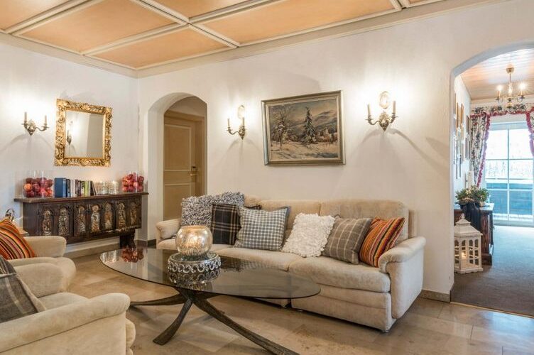 Ein Wohnzimmer mit beigen Sofas, einem Couchtisch aus Glas, gemusterten Kissen, Wandleuchten, gerahmten Kunstwerken und einer Anrichte mit Büchern und Dekorationsartikeln.