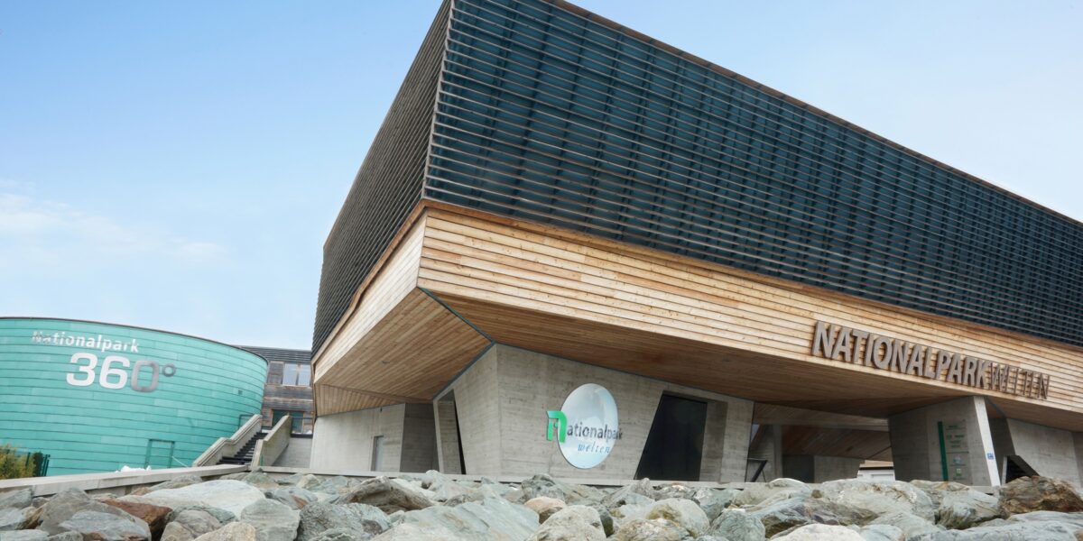 Ein modernes Gebäude mit Holz-, Glas- und Betontafeln mit der Aufschrift "Nationalpark" und "360°", mit großen Felsen im Vordergrund unter einem klaren blauen Himmel.