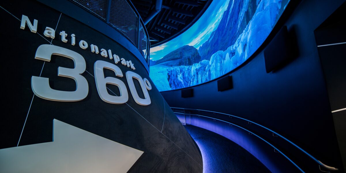 Geschwungener Gang mit "Nationalpark 360°"-Schildern an der Wand und einem großen Panoramadisplay, das eine Berglandschaft zeigt.