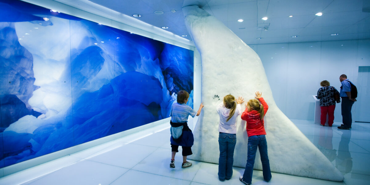 Drei Kinder berühren eine große Eisskulptur in einem hell erleuchteten, modernen Museumsraum mit einem blauen Gletscherfoto an der Wand; im Hintergrund sind drei Erwachsene zu sehen.