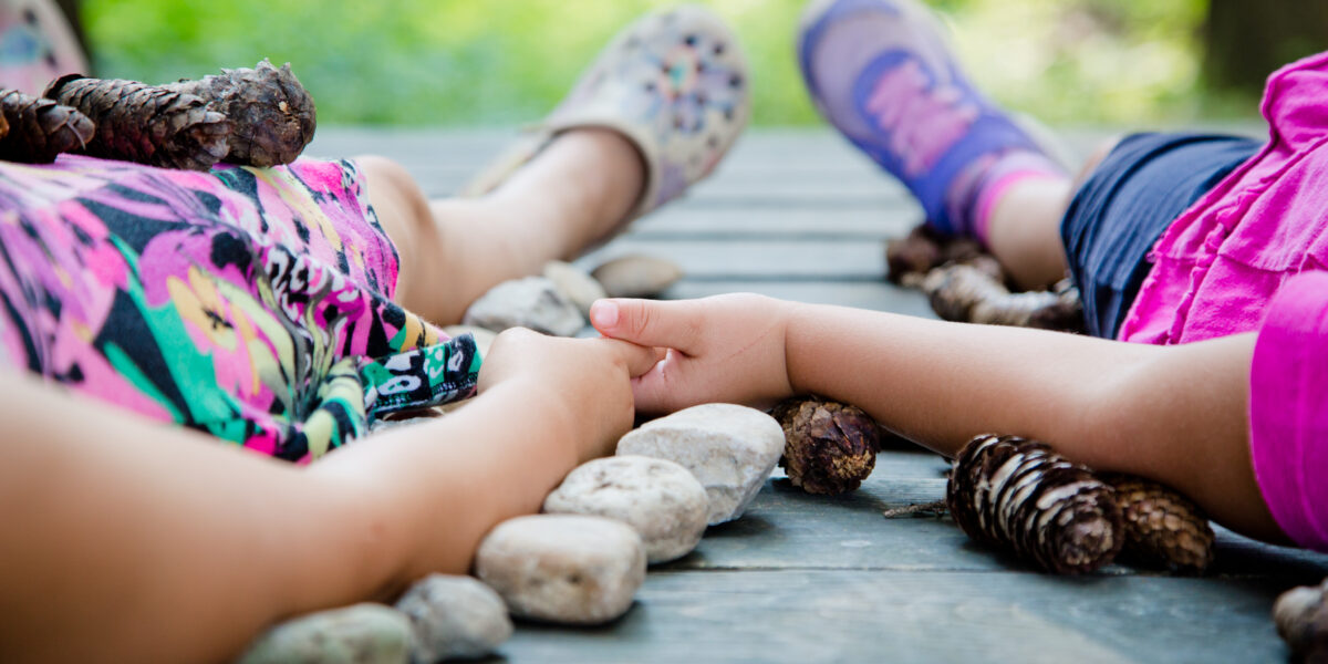 Zwei Kinder liegen händchenhaltend auf einer Holzfläche, umgeben von Tannenzapfen und Steinen. Ihre Gesichter sind nicht zu sehen.