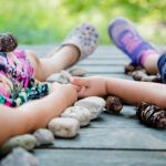 Zwei Kinder liegen händchenhaltend auf einer Holzfläche, umgeben von Tannenzapfen und Steinen. Ihre Gesichter sind nicht zu sehen.