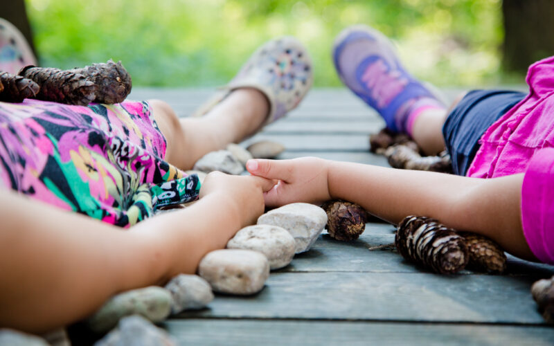 Zwei Kinder liegen händchenhaltend auf einer Holzfläche, umgeben von Tannenzapfen und Steinen. Ihre Gesichter sind nicht zu sehen.