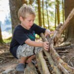 Kleiner Junge in schwarzem Hemd und Tarnshorts baut in einem Waldgebiet ein Gebäude aus Stöcken und Baumstämmen.