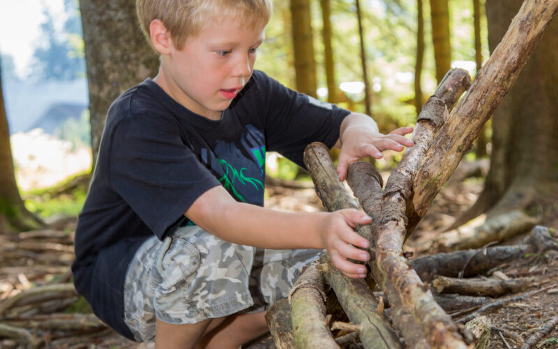 Kleiner Junge in schwarzem Hemd und Tarnshorts baut in einem Waldgebiet ein Gebäude aus Stöcken und Baumstämmen.