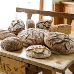 Auf einem rustikalen Holztisch in einem sonnendurchfluteten Raum mit Holzmöbeln liegen mehrere Brote mit verschiedenen Sorten aus.