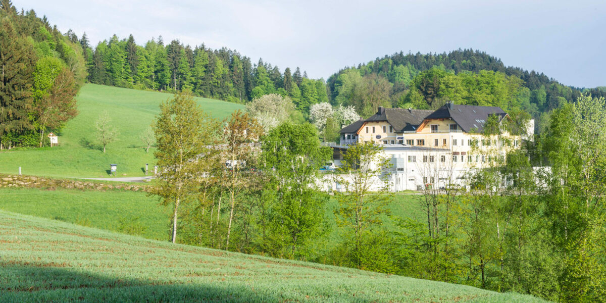Ein großes weißes Gebäude mit dunklen Dächern steht zwischen grünen Feldern und Bäumen, mit bewaldeten Hügeln im Hintergrund unter einem teilweise bewölkten Himmel.