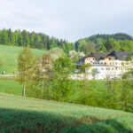 Ein großes weißes Gebäude mit dunklen Dächern steht zwischen grünen Feldern und Bäumen, mit bewaldeten Hügeln im Hintergrund unter einem teilweise bewölkten Himmel.