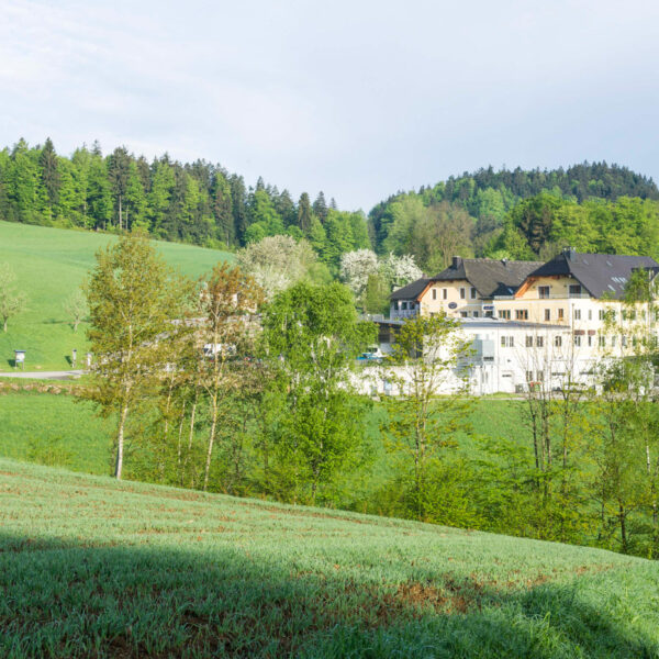 Ein großes weißes Gebäude mit dunklen Dächern steht zwischen grünen Feldern und Bäumen, mit bewaldeten Hügeln im Hintergrund unter einem teilweise bewölkten Himmel.