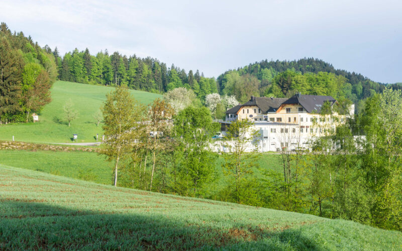 Ein großes weißes Gebäude mit dunklen Dächern steht zwischen grünen Feldern und Bäumen, mit bewaldeten Hügeln im Hintergrund unter einem teilweise bewölkten Himmel.