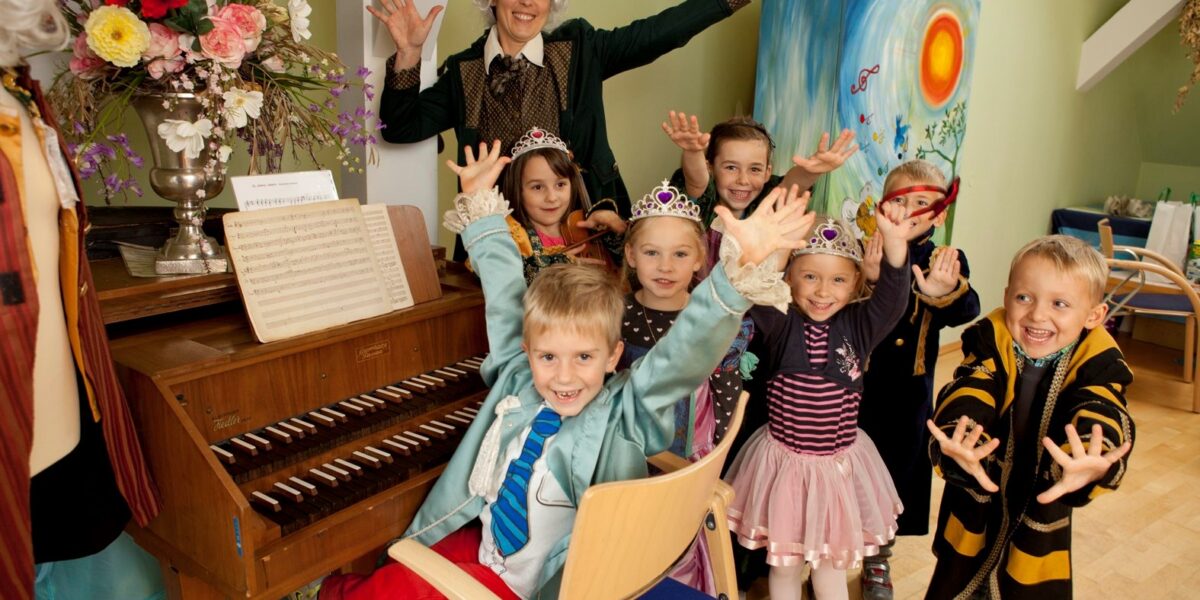 Eine Gruppe kleiner Kinder in Kostümen posiert mit einem Erwachsenen mit gepuderter Perücke neben einem Tasteninstrument in einem geschmückten Raum, alle lächeln mit erhobenen Händen.