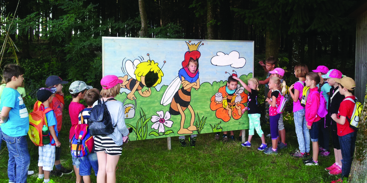 Eine Gruppe Kinder steht im Freien vor einer großen Tafel mit Comic-Bienen und Blumen und hört einer Frau zu, die teilweise als Biene verkleidet ist.