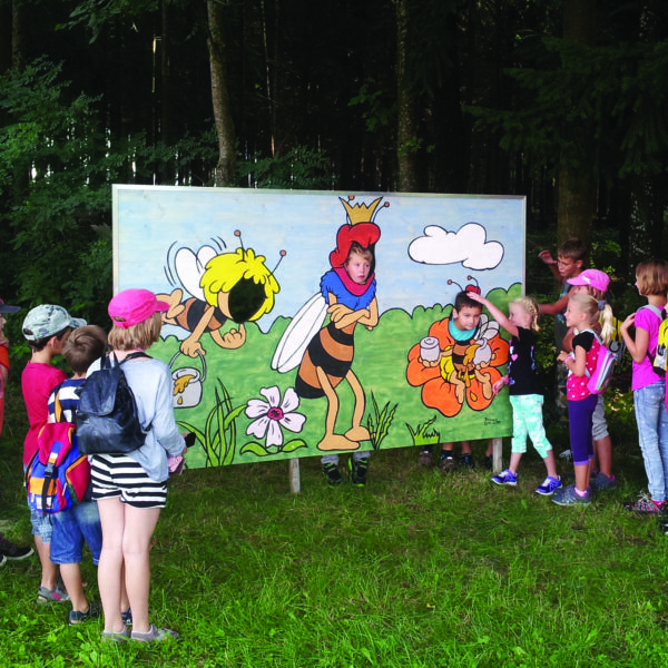 Eine Gruppe Kinder steht im Freien vor einer großen Tafel mit Comic-Bienen und Blumen und hört einer Frau zu, die teilweise als Biene verkleidet ist.