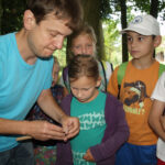 Ein Erwachsener steht im Freien zwischen Bäumen und zeigt einer Gruppe von Kindern einen Gegenstand, den er in der Hand hält. Die Kinder schauen aufmerksam zu.