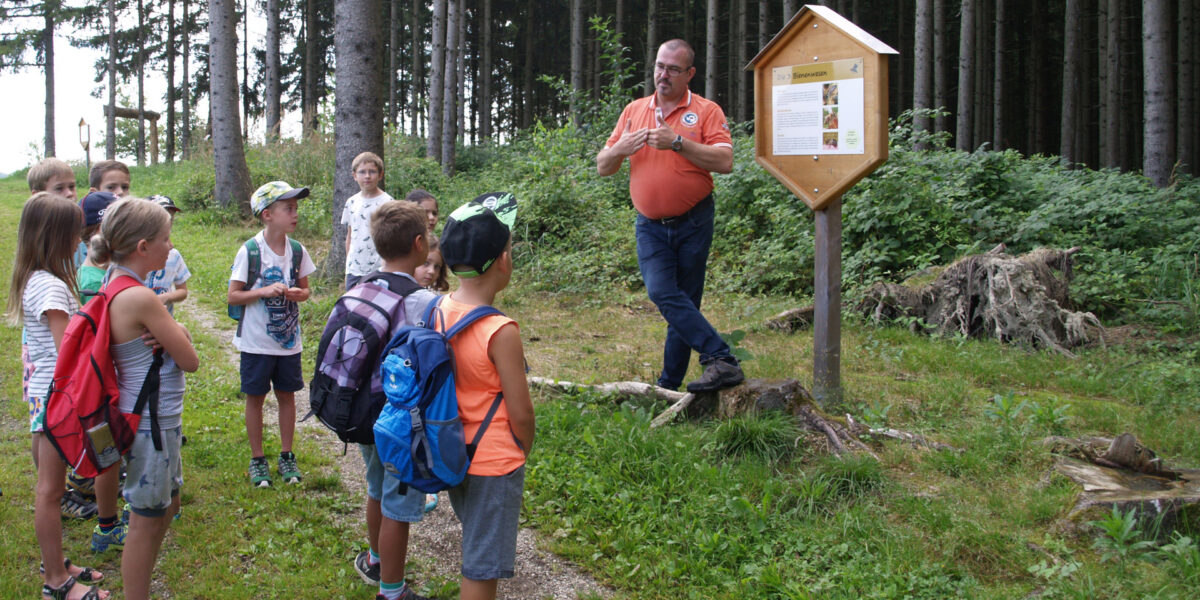 Ein Mann hält einer Gruppe von Kindern neben einem Informationsschild in einem Waldstück einen informativen Vortrag. Die Kinder tragen Rucksäcke und hören aufmerksam zu.