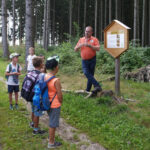 Ein Mann hält einer Gruppe von Kindern neben einem Informationsschild in einem Waldstück einen informativen Vortrag. Die Kinder tragen Rucksäcke und hören aufmerksam zu.