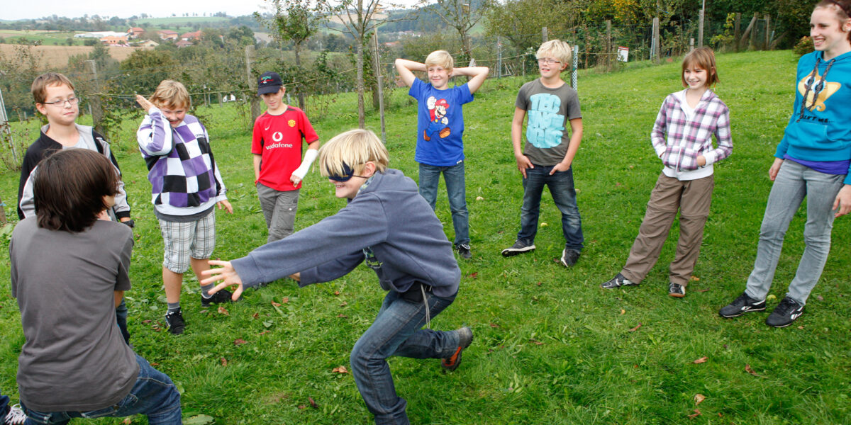 Eine Gruppe Kinder steht an einem bewölkten Tag in einem Kreis auf dem Rasen und beobachtet zwei Kinder, die in der Mitte ein Spiel spielen.