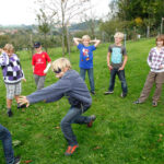 Eine Gruppe Kinder steht an einem bewölkten Tag in einem Kreis auf dem Rasen und beobachtet zwei Kinder, die in der Mitte ein Spiel spielen.